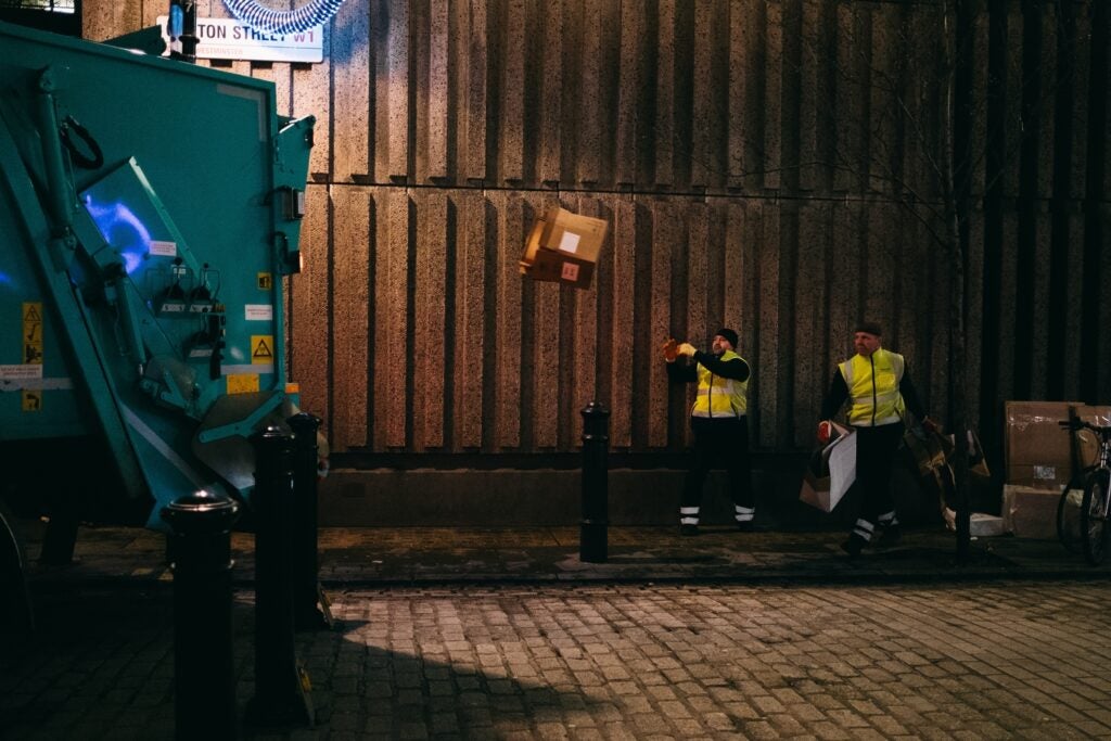 two sanitation workers throwing trash into the back of a garbage truck