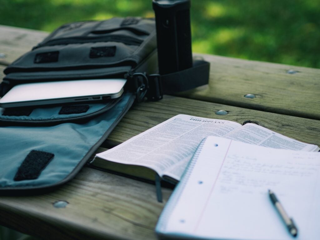 a backpack with a laptop in it on a wooden table outside, with an open book and a notebook and pen