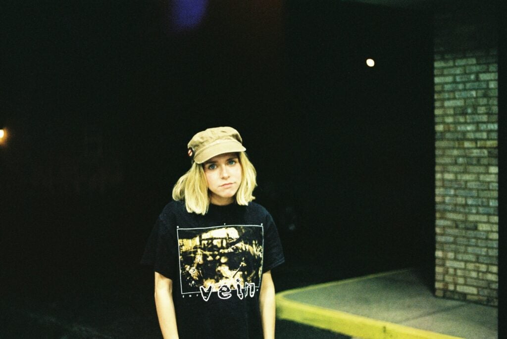 a blonde woman in a black t-shirt standing outside a building in a parking lot at night