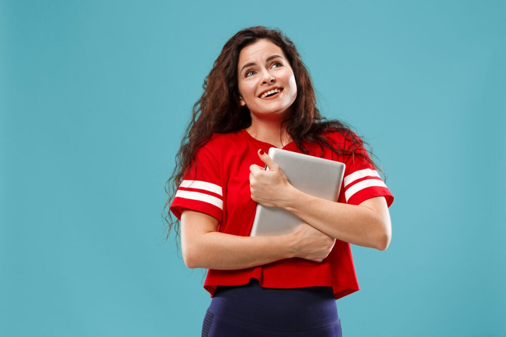 A brunette woman wearing a red short-sleeved shirt and hugging a silver laptop in front of a solid blue background.