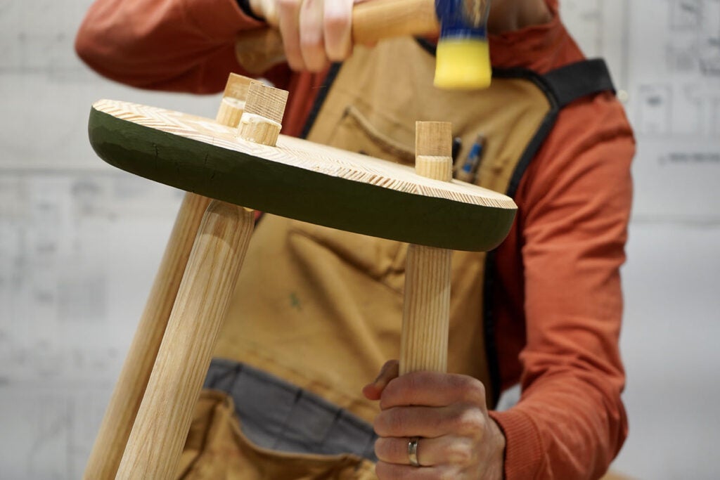 a person using a mallet to drive wooden wedges into a slot in the tenon of the legs of a wooden three-legged stool