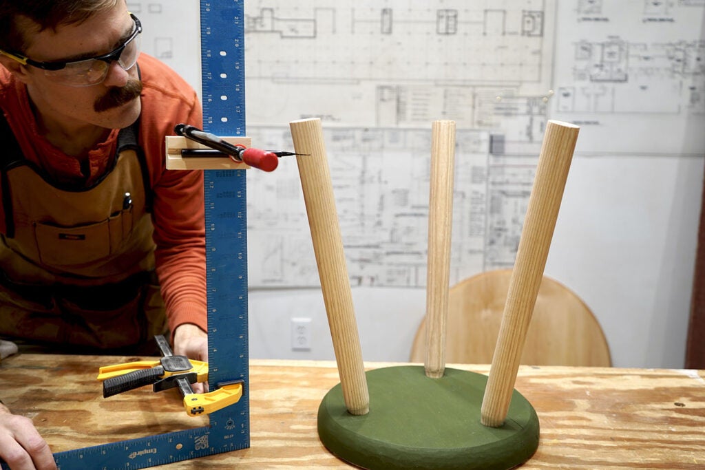 a person using a carpenter's square, a pencil, and a smaller square to ensure they draw level lines on the legs of a wooden three-legged stool