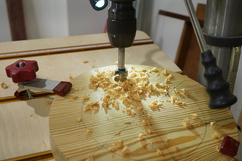 a pine stool seat under a drill press as a person bores holes for legs