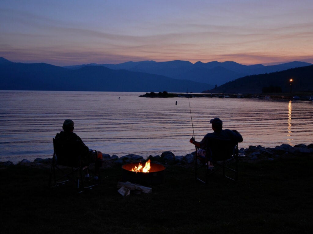 two people sitting by the water, burning a campfire and fishing at sunset