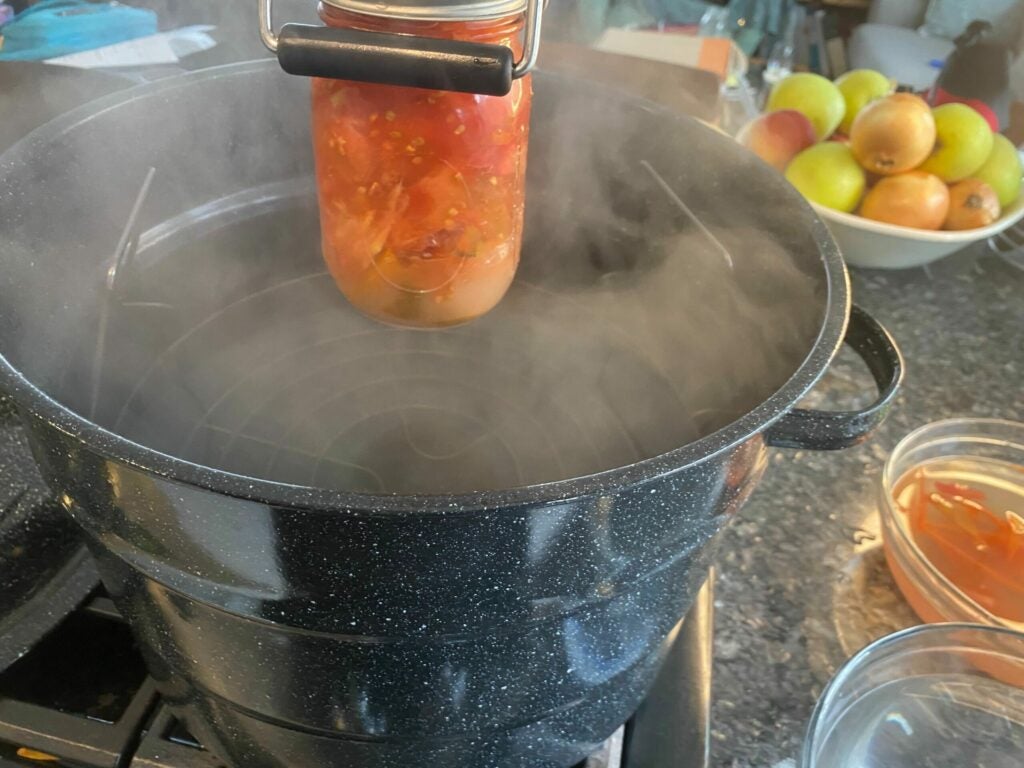 a person removing a jar of canned tomatoes from a pot of boiling water with a jar lifter